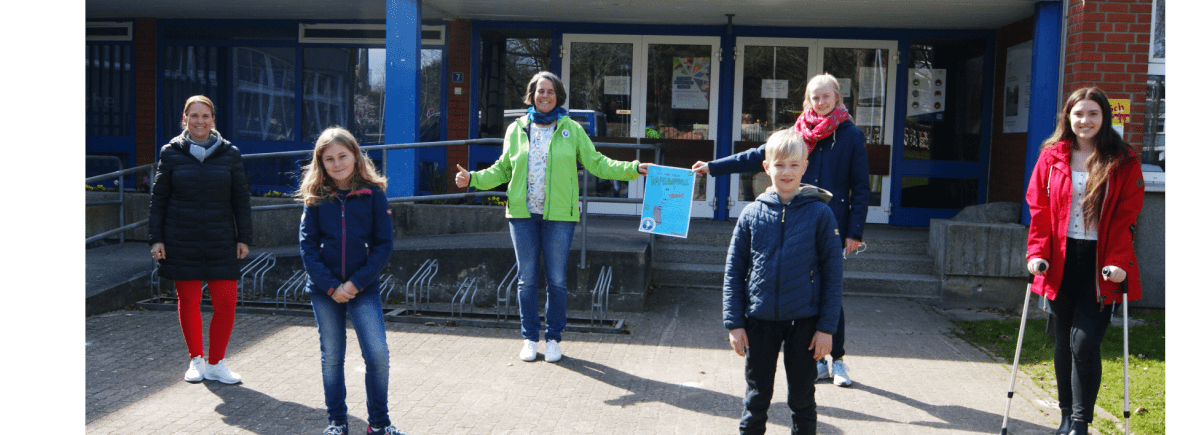 Vier Frauen und zwei Kinder stehen vor einer Schule und halten ein Plakat für einen Schülermalwettbewerb hoch